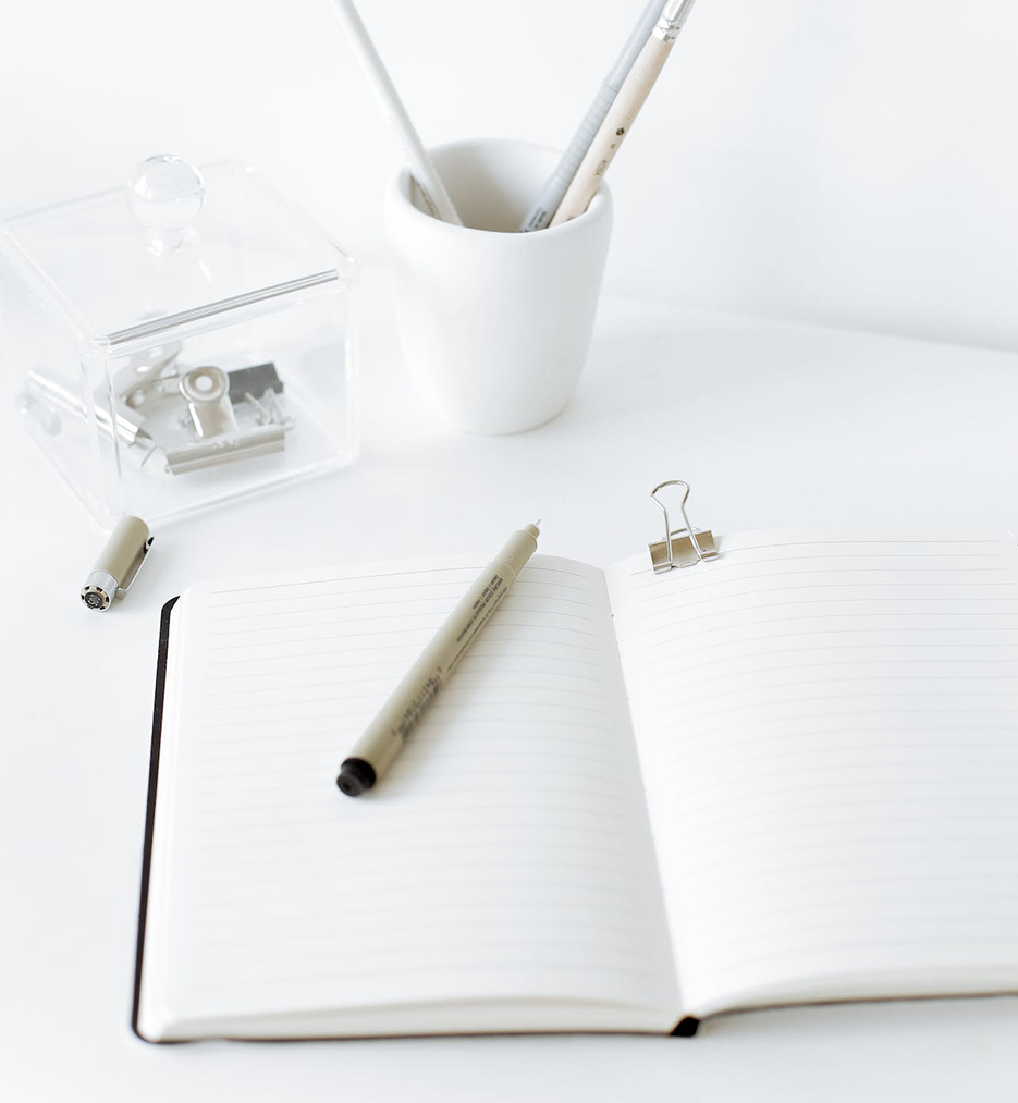 A notebook opened to an empty page with a pen on top and stationary behind