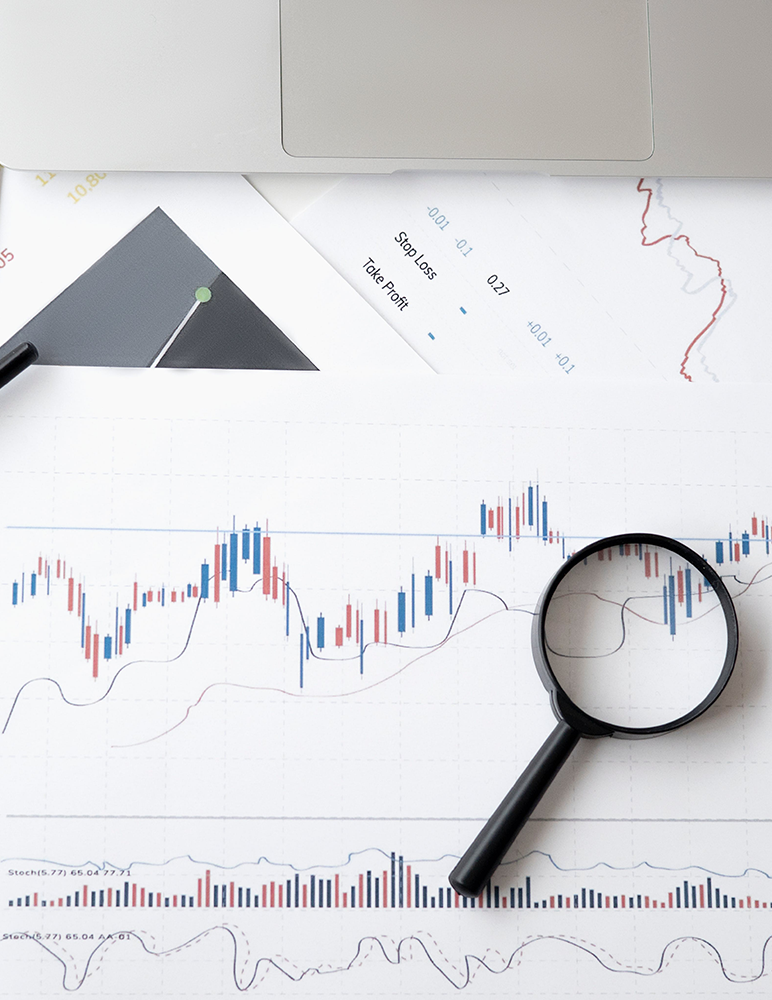 Some printed charts and market trading details with a magnifying glass on top, and a MacBook Air trackpad visible on top