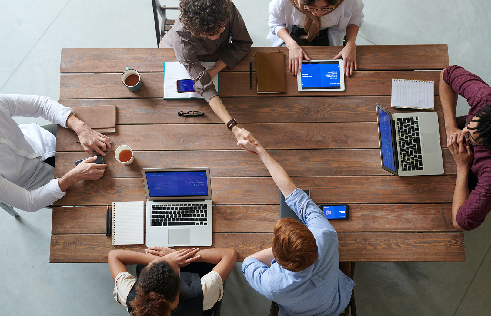 Several people sitting at a table with laptops, books and drinks, two people shaking hands across the table